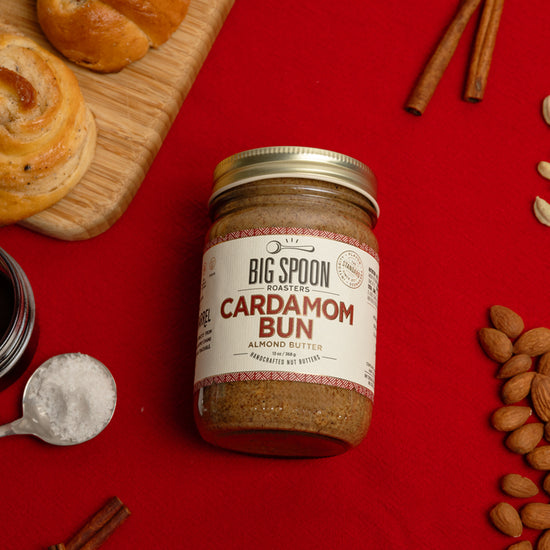 jar of Cardamom Bun Almond Butter on a red tablecloth surrounded by cardamom buns on a wooden board, cinnamon sticks, a spoonful of salt, a jar of coconut nectar, vanilla beans, cardamom pods, and almonds.
