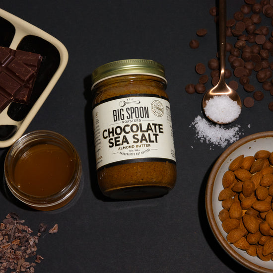 a jar of Chocolate Sea Salt almond butter surrounded by a dish of chocolate bars, a jar of honey, a spoonful of sea salt, chocolate chips, almonds, and cacao nibs