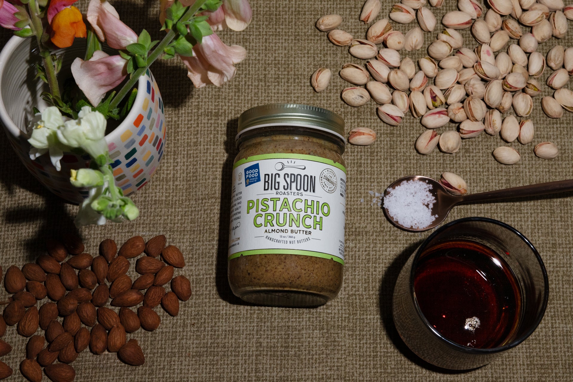 A jar of Pistachio Crunch Almond Butter surrounded by almonds, pistachios, a spoonful of sea salt, and a jar of maple syrup, alongside a vase of flowers