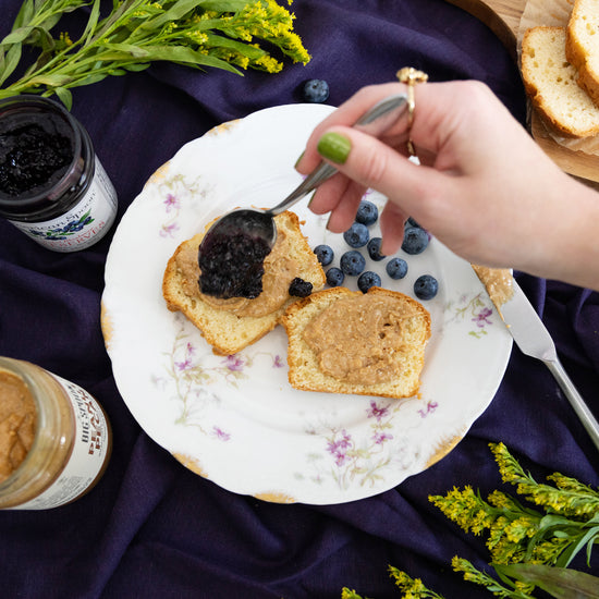 Hand drizzling blueberry jam over 2 pieces of toast with peanut pecan nut butter spread on a plate with berries. Below the plate is an indigo table cloth.
