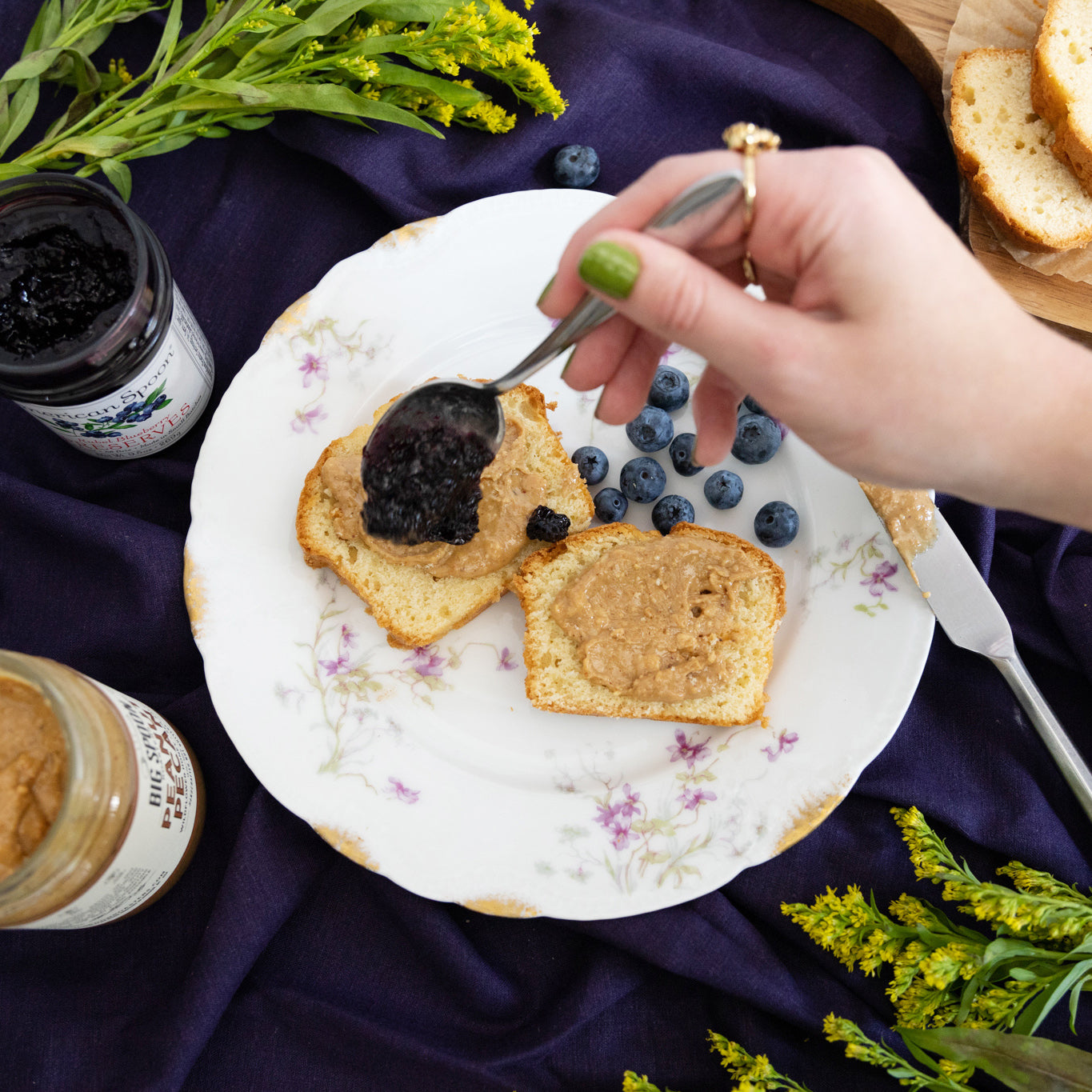 Hand drizzling blueberry jam over 2 pieces of toast with peanut pecan nut butter spread on a plate with berries. Below the plate is an indigo table cloth.