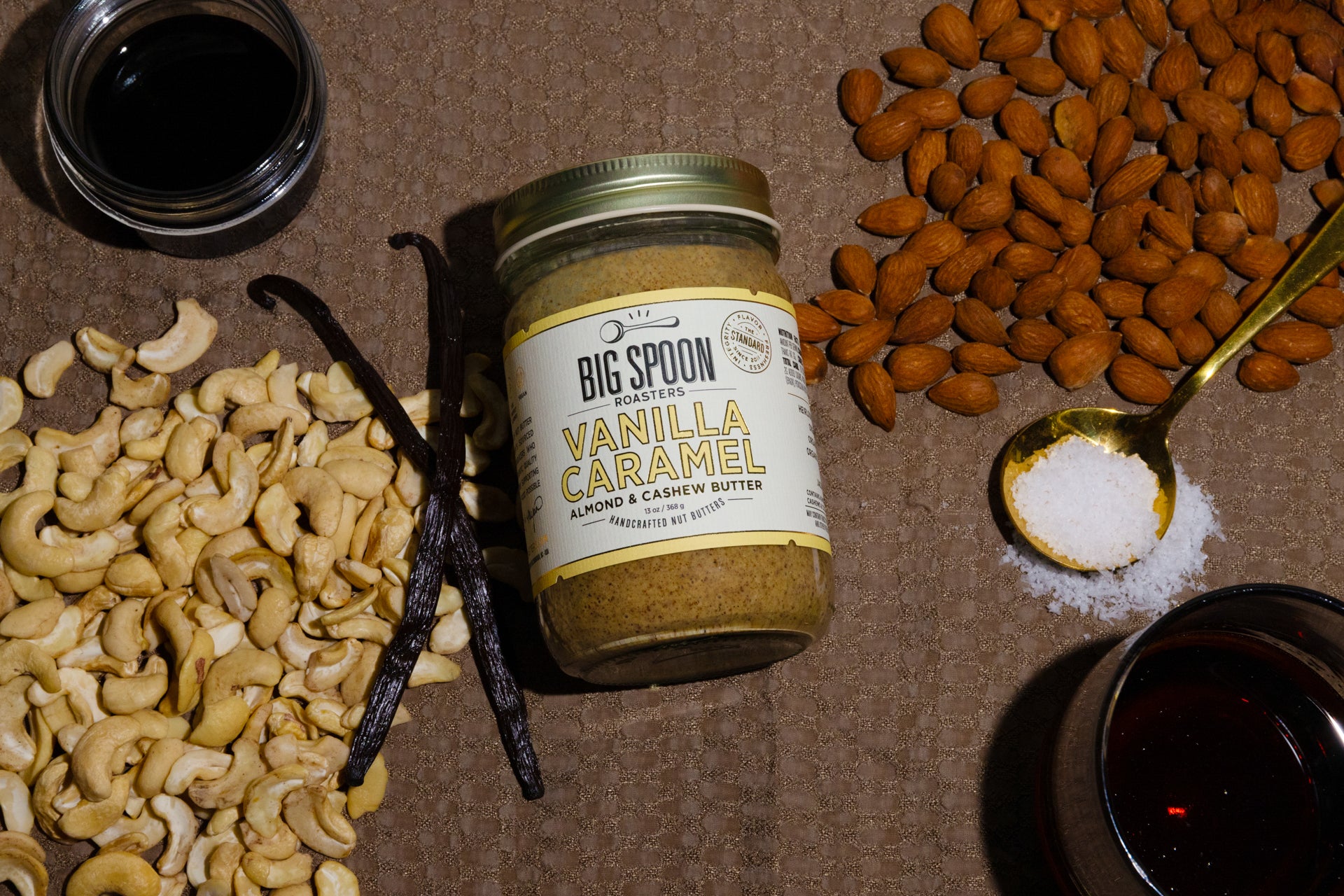 Vanilla Caramel Almond & Cashew Butter on a brown background surrounded by almonds, cashews, vanilla bean,  spoonful of sea salt, and open jars of coconut nectar and maple syrup