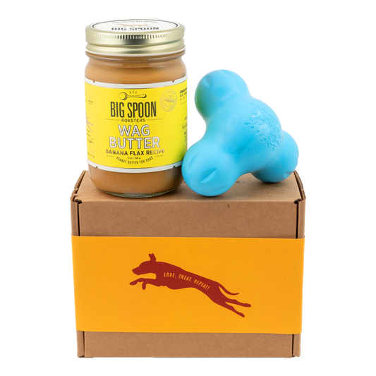 Jar of Banana Flax Wag Butter and a blue West Paw Tux toy sit on top of a gift box with a gold and maroon decorative dog label.