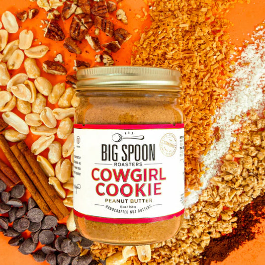 13oz glass jar of Cowgirl Cookie Peanut Butter with coconut flakes, pecan pieces, cashews, cinnamon sticks, chocolate chips, sea salt, granola, and vanilla powder spread out on an orange background.