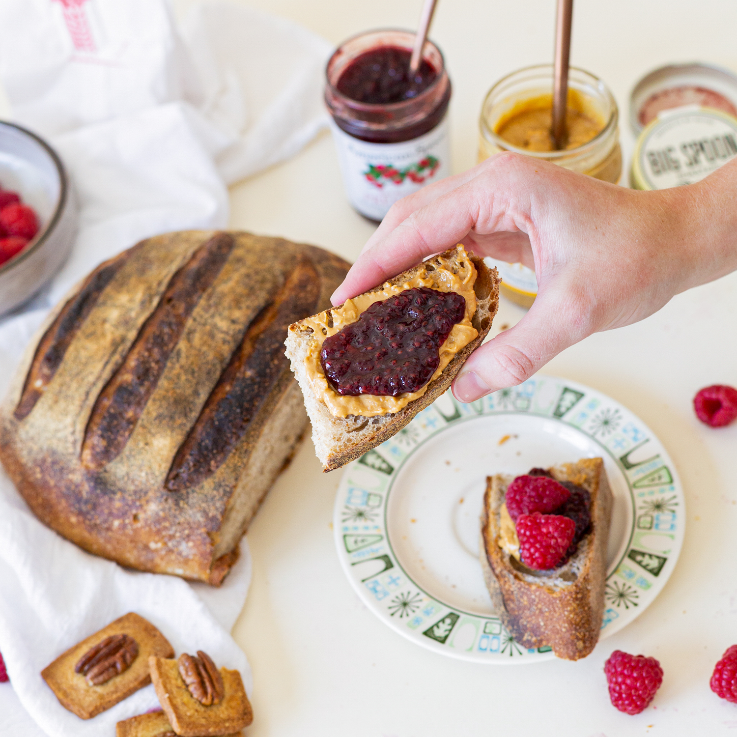 A hand holds a piece of bread with peanut butter and jam in front of a spread of jam, peanut butter, bread, and fresh fruit