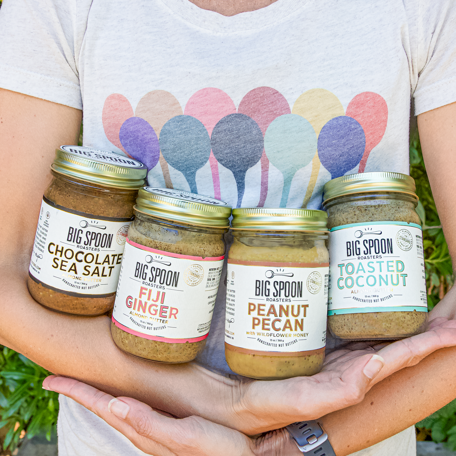 A person wearing a colorful spoons t-shirt holds jars of Chocolate Sea Salt, Fiji Ginger, Peanut Pecan, and Toasted Coconut