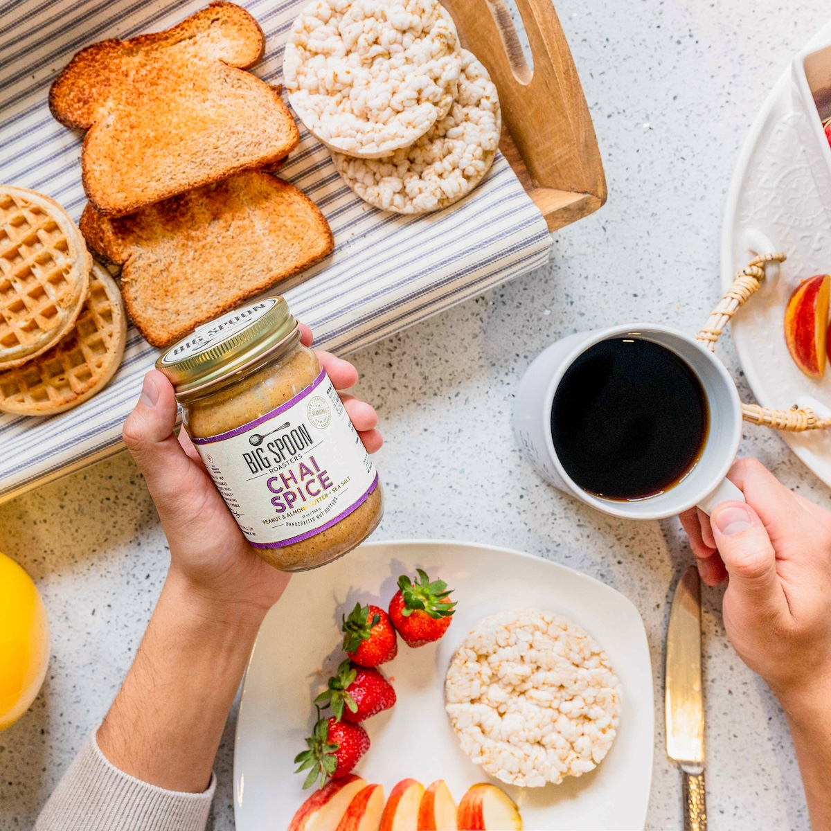 A hand holds a jar of Chai Spice nut butter surrounded by breakfast options like toast, waffles, and rice cakes along with a cup of black coffee