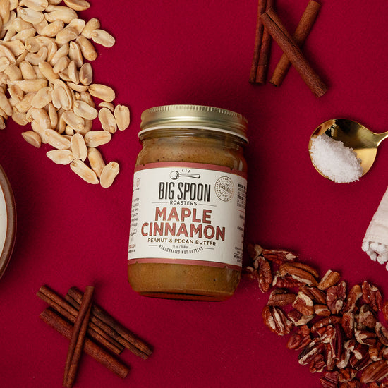 Atop a red background, a jar of Maple Cinnamon Peanut & Pecan Butter is surrounded by peanuts, pecans, cinnamon sticks, a bowl of maple syrup, and a spoonful of sea salt on top of a cloth napkin.