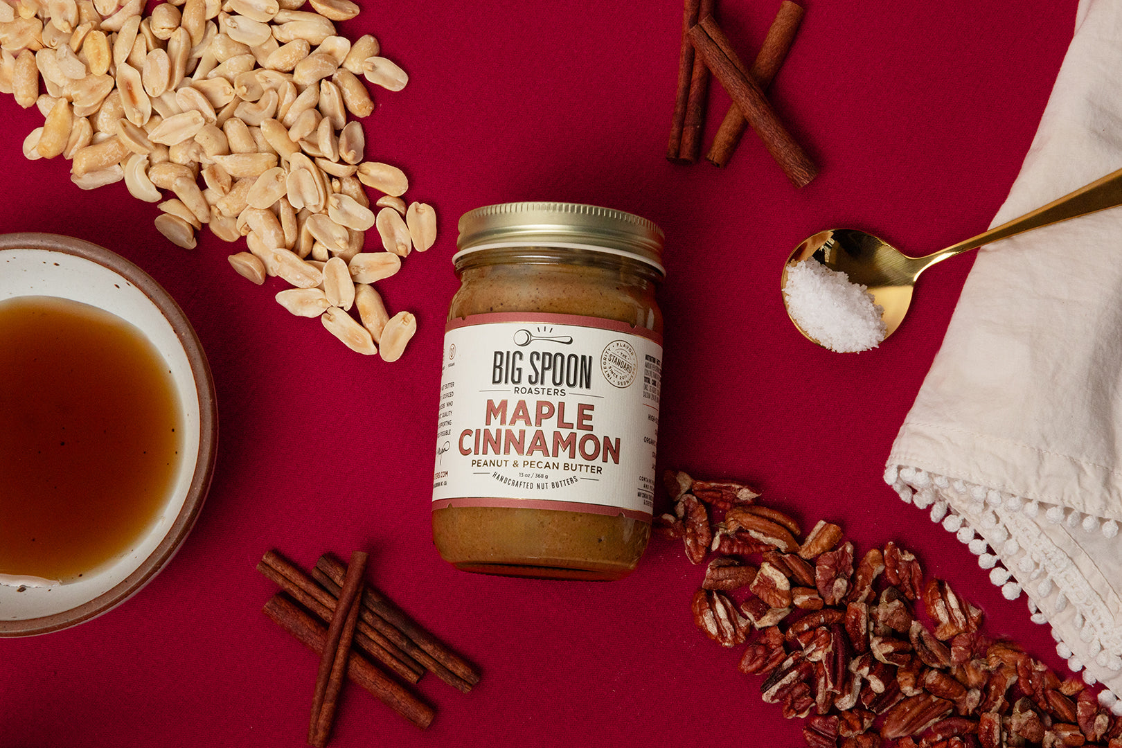 Atop a red background, a jar of Maple Cinnamon Peanut & Pecan Butter is surrounded by peanuts, pecans, cinnamon sticks, a bowl of maple syrup, and a spoonful of sea salt on top of a cloth napkin.