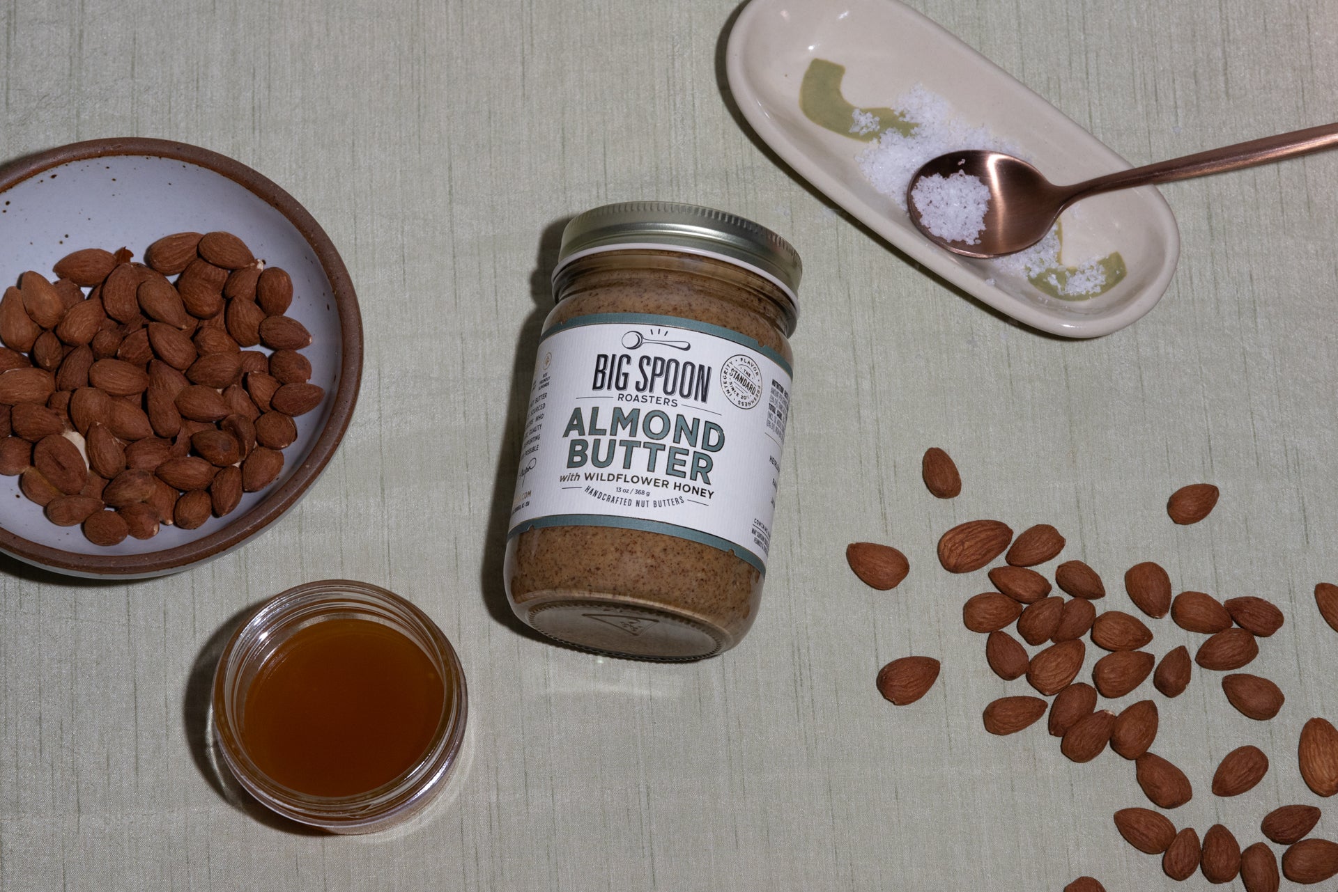 a jar of Almond Butter surrounded by a bowl of almonds, a jar of honey, sea salt, and other almonds