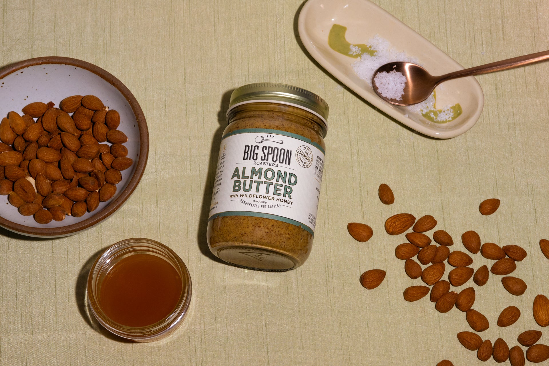 a jar of Almond Butter surrounded by a bowl of almonds, a jar of honey, sea salt, and other almonds