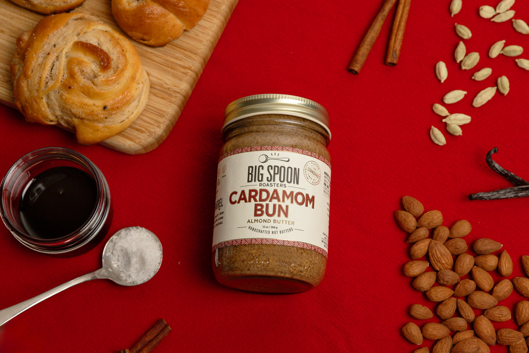 jar of Cardamom Bun Almond Butter on a red tablecloth surrounded by cardamom buns on a wooden board, cinnamon sticks, a spoonful of salt, a jar of coconut nectar, vanilla beans, cardamom pods, and almonds.