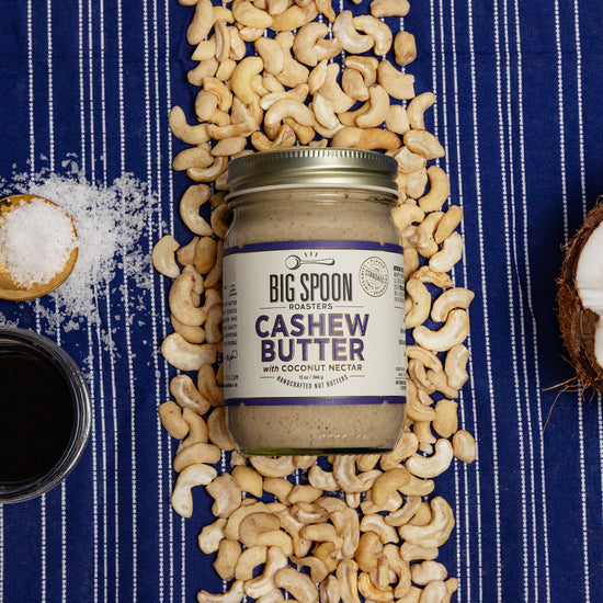 Jar of Big Spoon Cashew Butter surrounded by cashews, coconut, coconut nectar and oil, and salt on a blue striped background