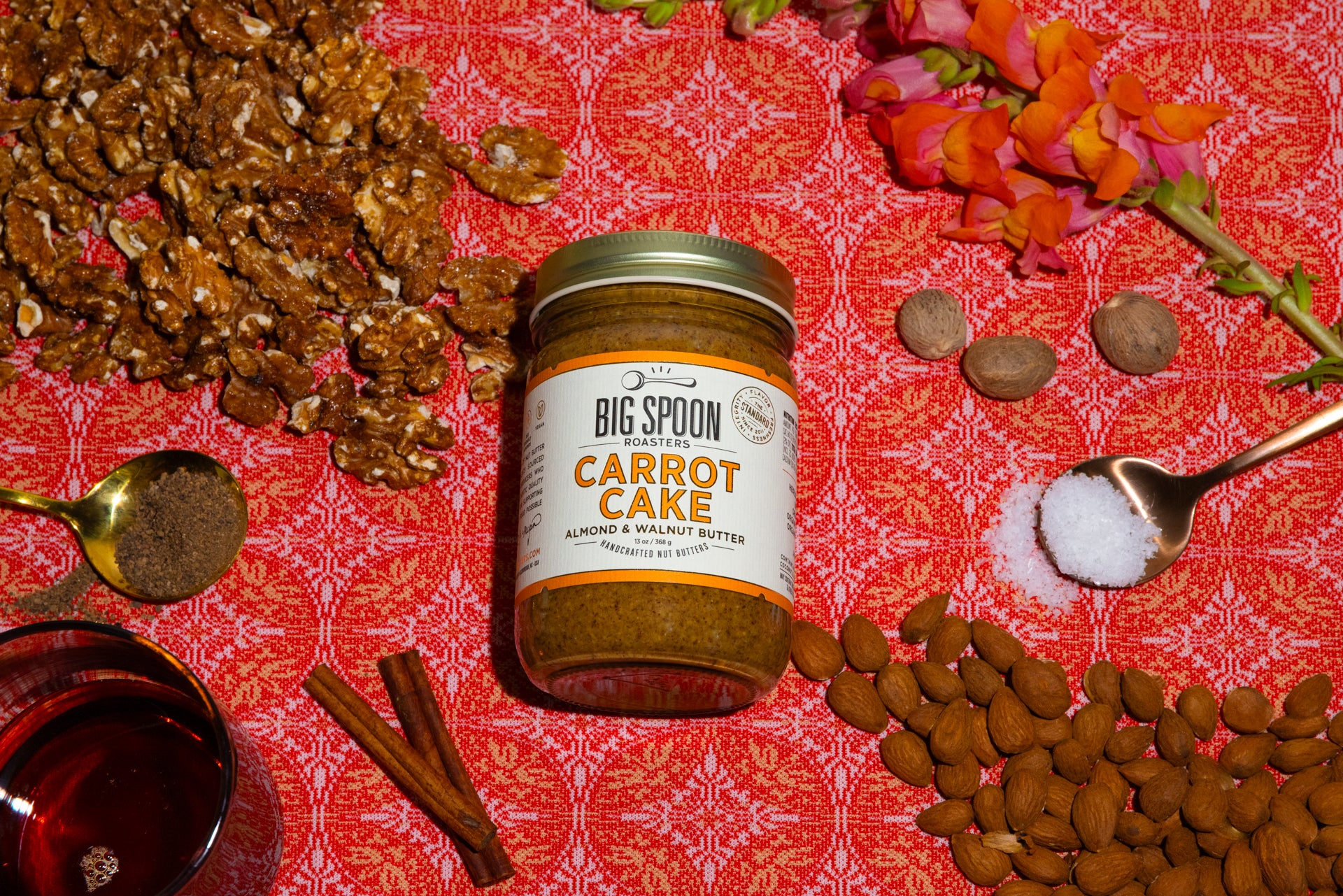 a jar Carrot Cake Almond & Walnut Butter surrounded by walnuts, a spoonful of spices, a jar of maple syrup, cinnamon sticks, whole nutmeg, a spoonful of sea salt, and almonds