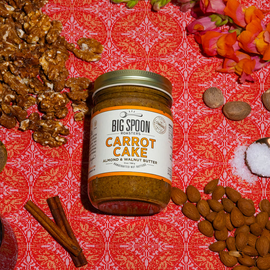 a jar Carrot Cake Almond & Walnut Butter surrounded by walnuts, a spoonful of spices, a jar of maple syrup, cinnamon sticks, whole nutmeg, a spoonful of sea salt, and almonds