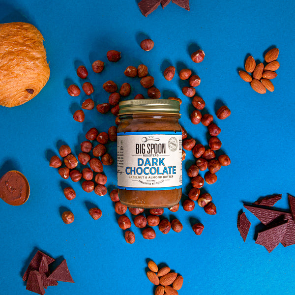 ALT TEXT: A 13 oz glass jar of Dark Chocolate Hazelnut & Almond Butter on a deep blue background surrounded by hazelnuts, almonds, dark chocolate, a golden spoon with nut butter, and a dark chocolate pain au chocolat.