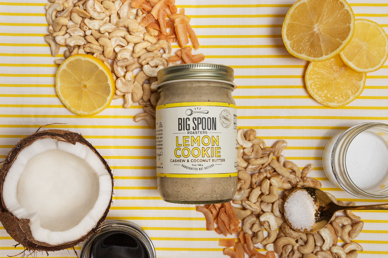 Jar of Lemon Cookie Cashew Butter with lemon, coconut, cashews, salt, and coconut nectar/oil around it on a yellow striped background