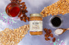 Jar of Big Spoon Toffee Crunch surrounded by cashews, pecans, peanuts, salt, and small bowls of syrup on a metallic background.