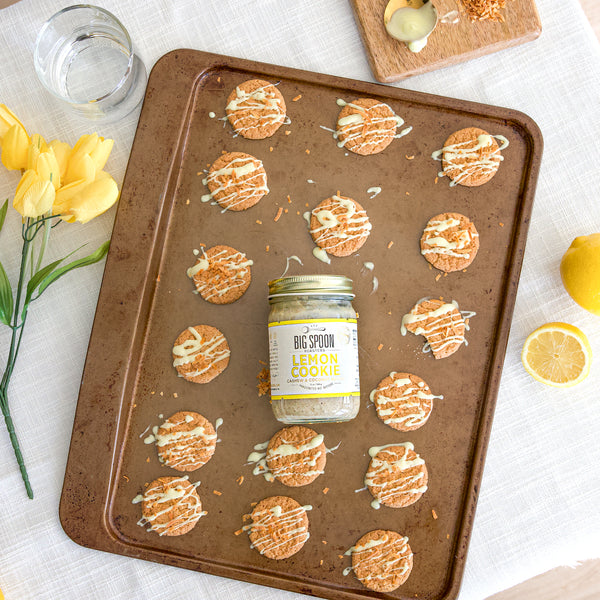 13oz jar of Lemon Cookie Cashew Butter on a cookie sheet surrounded by lemon cookies on a light linen cloth table surrounded by a fresh cut lemon, a cutting board, a glass of water, and a small bunch of yellow tulips.