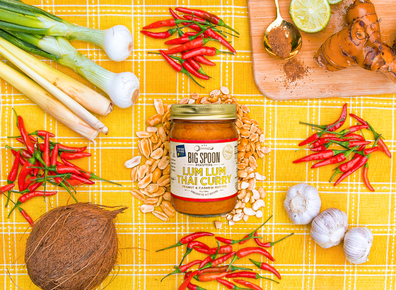 13oz Lum Lum Thai Curry Peanut & Cashew Butter surrounded by red chilis, peanuts, cashews, shallots, coconut and green onion. A cutting board topped with a golden spoon, coconut sugar, limes and ginger all sits on a yellow gingham cloth. 