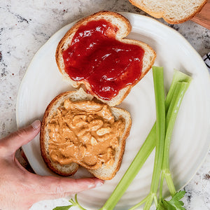 A hand reaches for a piece of peanut butter toast with a toast with jam and celery sticks laying next to it on a white plate and marble countertop