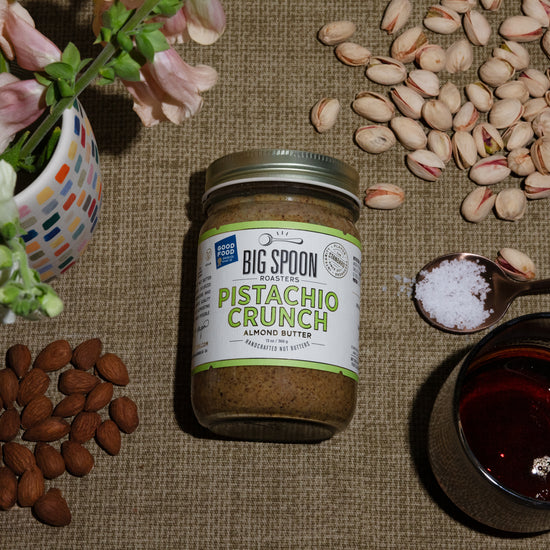 A jar of Pistachio Crunch Almond Butter surrounded by almonds, pistachios, a spoonful of sea salt, and a jar of maple syrup, alongside a vase of flowers