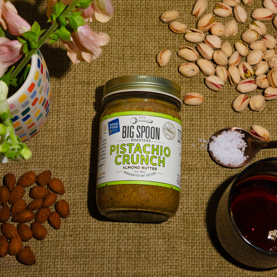 A jar of Pistachio Crunch Almond Butter surrounded by almonds, pistachios, a spoonful of sea salt, and a jar of maple syrup, alongside a vase of flowers