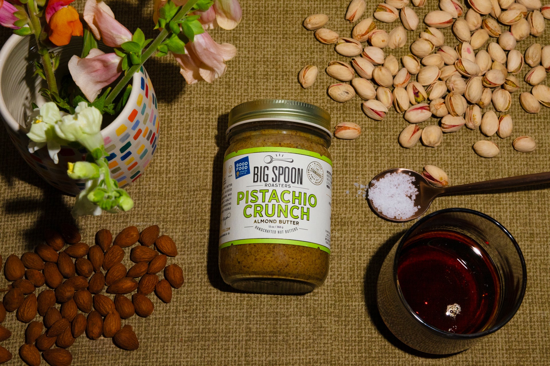 A jar of Pistachio Crunch Almond Butter surrounded by almonds, pistachios, a spoonful of sea salt, and a jar of maple syrup, alongside a vase of flowers