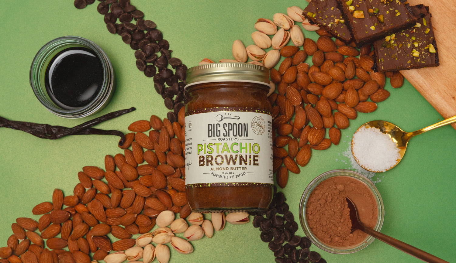 Jar of Big Spoon Pistachio Brownie Nut Butter surrounded by almonds, pistachios, chocolate chips, vanilla, coconut nectar, cocoa powder, a spoonful of salt, and pistachio-topped brownies on a green background.