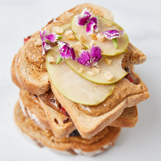 Several pieces of toast stacked with almond butter, pear slices, fiji ginger bits, and purple edible flowers