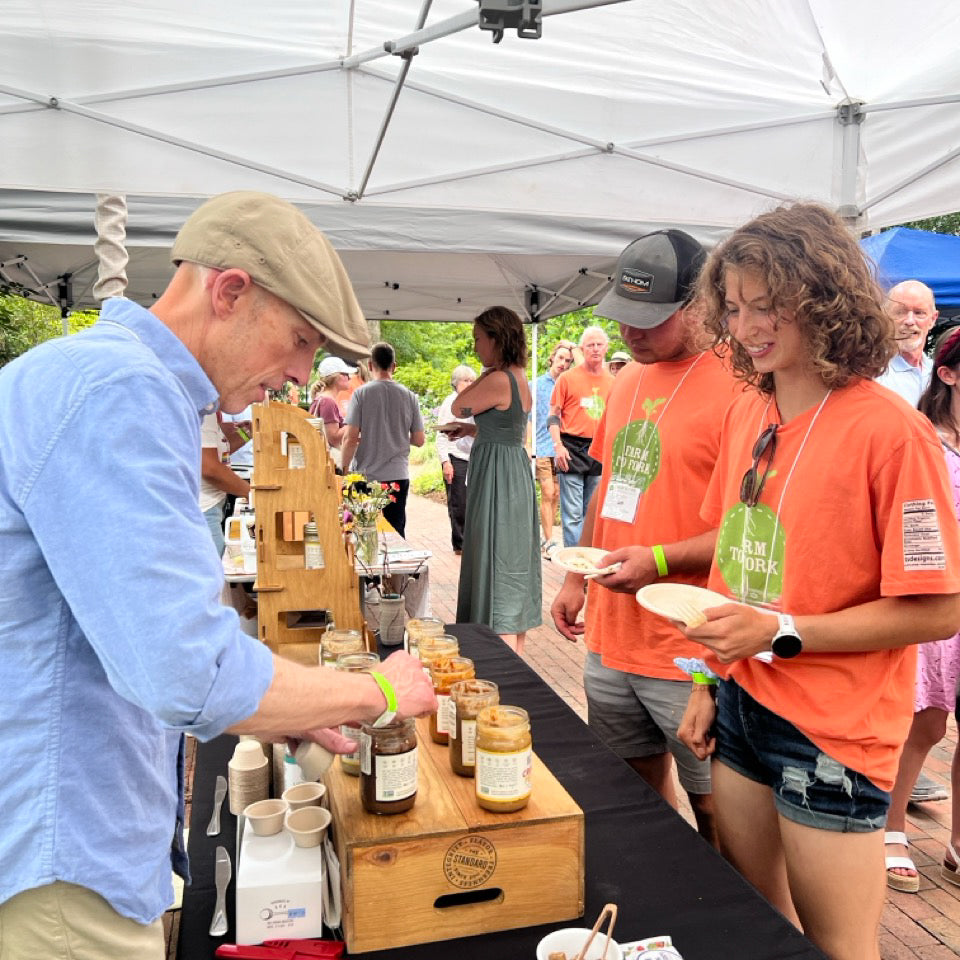 A man in an blue shirt and tan hat scoops nut butter out of a jar at a tasting event to hand to a woman in an orange shirt smiling