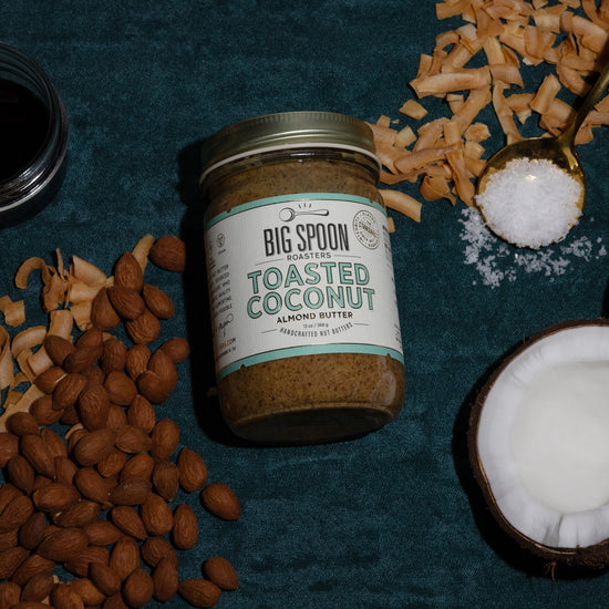 A jar of Toasted Coconut Almond Butter sits alongside almonds, toasted coconut chips, a jar of coconut nectar, a spoonful of sea salt, and a whole opened coconut