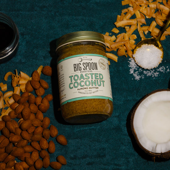 A jar of Toasted Coconut Almond Butter sits alongside almonds, toasted coconut chips, a jar of coconut nectar, a spoonful of sea salt, and a whole opened coconut