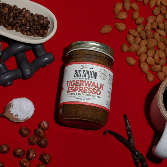 A jar of Tigerwalk Espresso Almond & Hazelnut Butter sits alongside coffee beans, hazelnuts, a jar of coconut nectar, a spoonful of sea salt, almonds, vanilla bean, and a mug of coffee