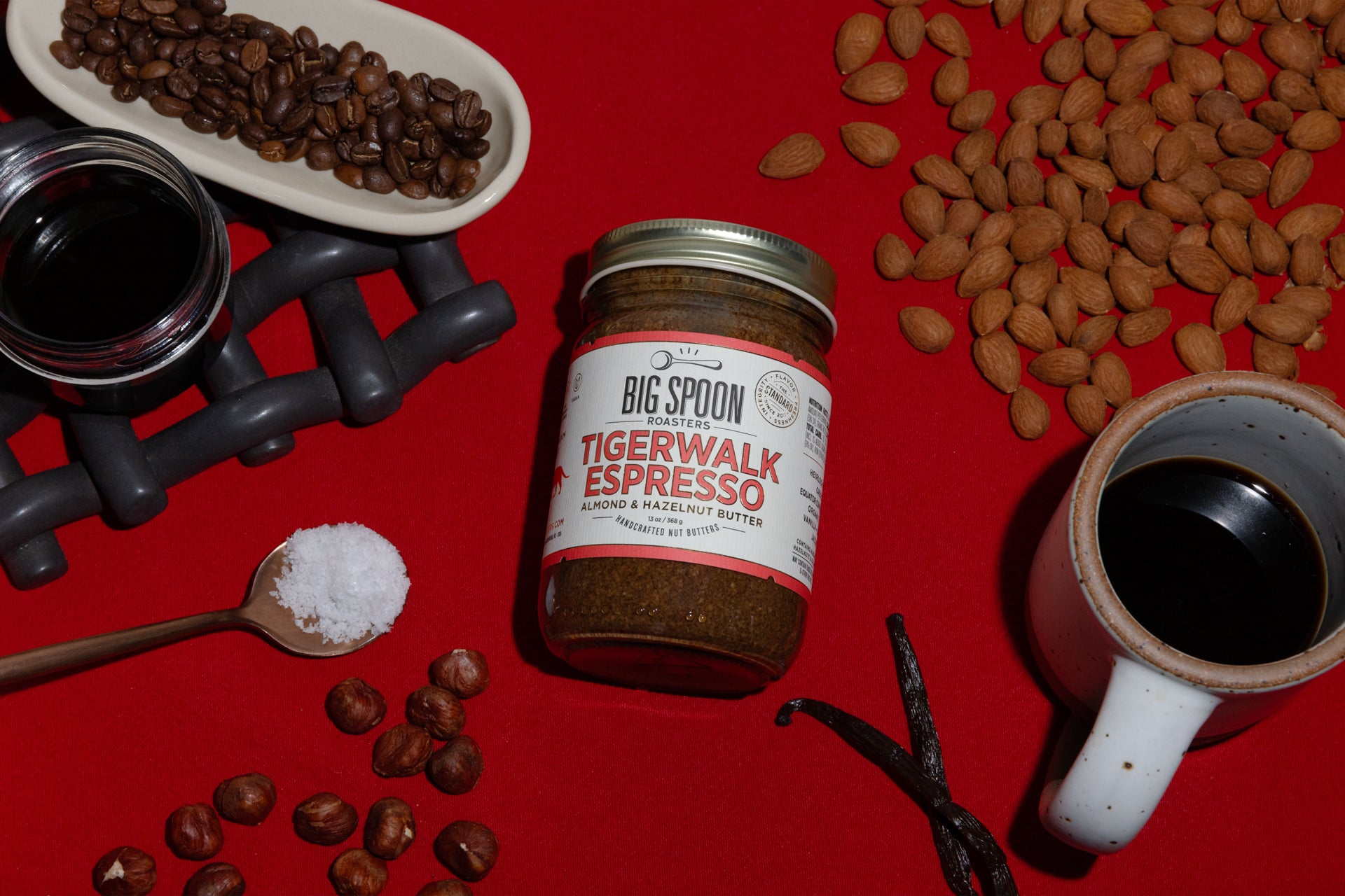 A jar of Tigerwalk Espresso Almond & Hazelnut Butter sits alongside coffee beans, hazelnuts, a jar of coconut nectar, a spoonful of sea salt, almonds, vanilla bean, and a mug of coffee