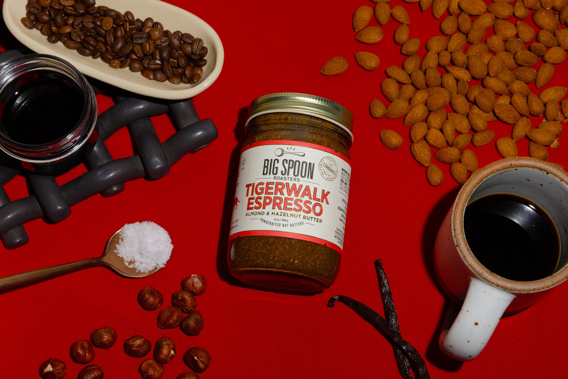 A jar of Tigerwalk Espresso Almond & Hazelnut Butter sits alongside coffee beans, hazelnuts, a jar of coconut nectar, a spoonful of sea salt, almonds, vanilla bean, and a mug of coffee