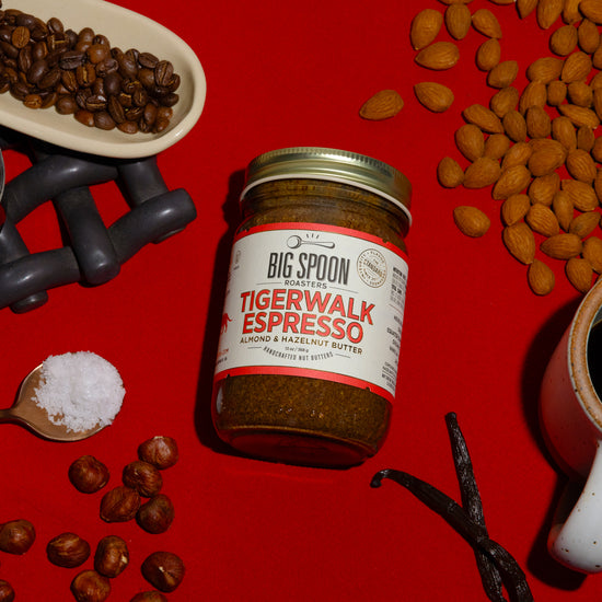 A jar of Tigerwalk Espresso Almond & Hazelnut Butter sits alongside coffee beans, hazelnuts, a jar of coconut nectar, a spoonful of sea salt, almonds, vanilla bean, and a mug of coffee