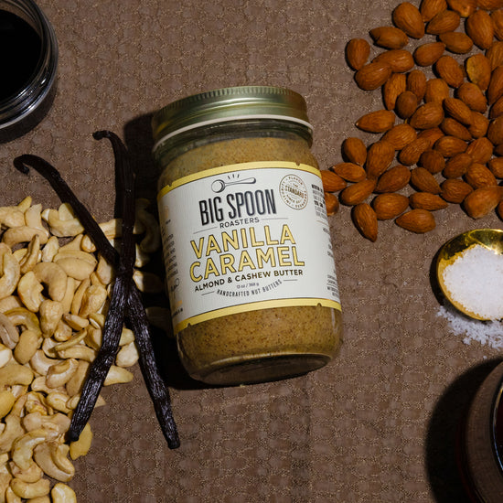 Vanilla Caramel Almond & Cashew Butter on a brown background surrounded by almonds, cashews, vanilla bean,  spoonful of sea salt, and open jars of coconut nectar and maple syrup