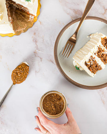 A hand reaches for a jar of Carrot Cake nut butter surrounded by a whole cake, a slice of carrot cake, and a spoon of Carrot Cake nut butter on top of a light marble countertop