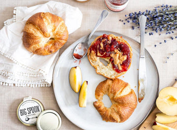 A croissant is split in half with jam, peanut butter spread on one side, surrounded by fresh fruit, lavender, a croissant, and a white cloth napkin