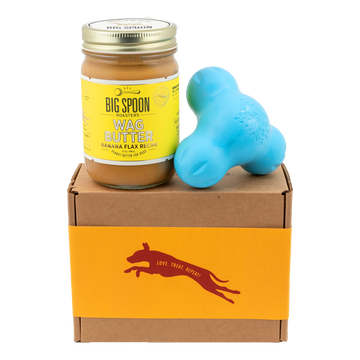 Jar of Banana Flax Wag Butter and a blue West Paw Tux toy sit on top of a gift box with a gold and maroon decorative dog label.