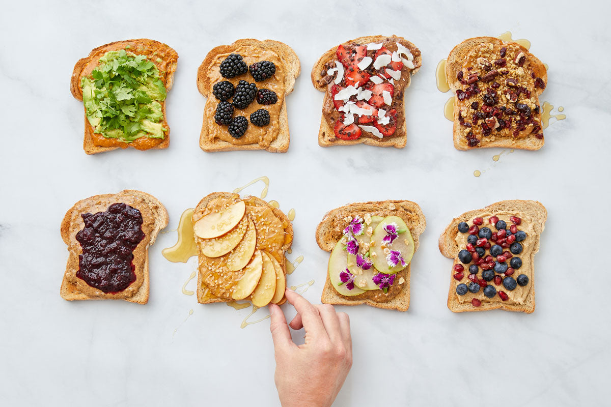 Eight different nut butter toasts with various fruit and decorative toppings, hand reaching for one